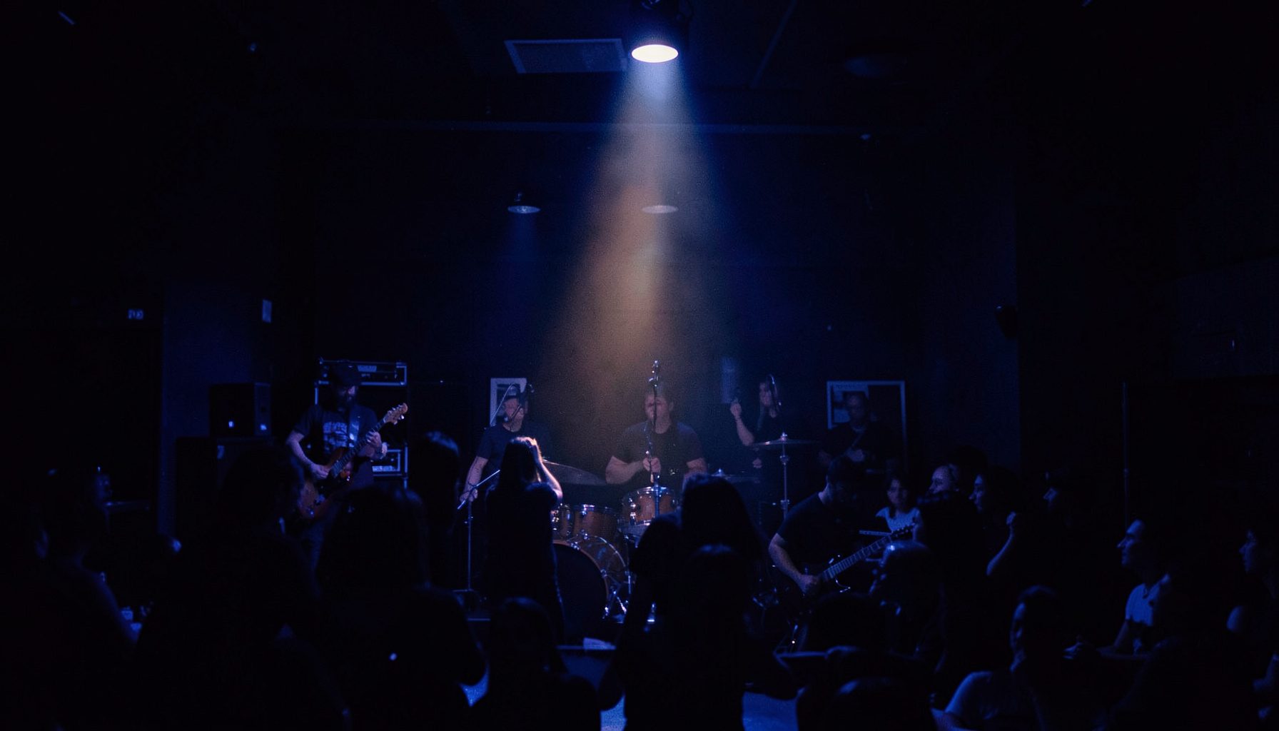 A moody, dimly lit live-music venue interior with deep indigo shadows and a single warm spotlight glow