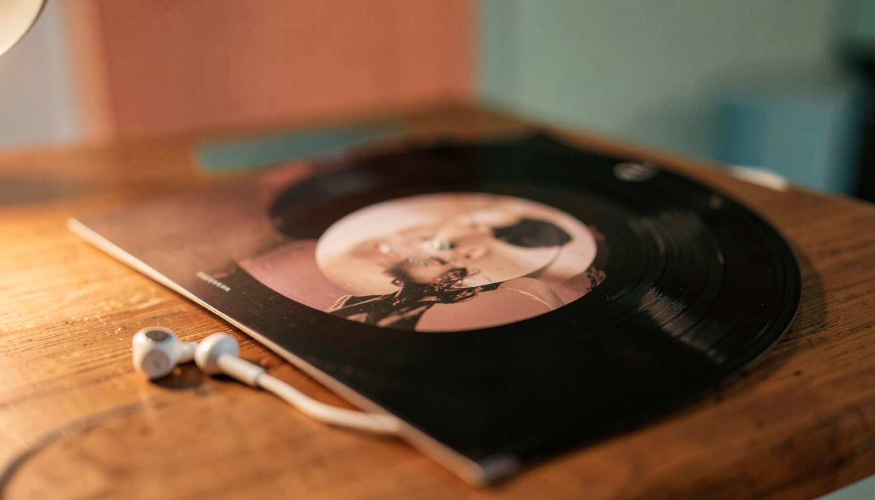 A warm, softly lit close-up of a vinyl record sleeve and a pair of earbuds resting on a wooden table, with muted coral and teal tones in the background.