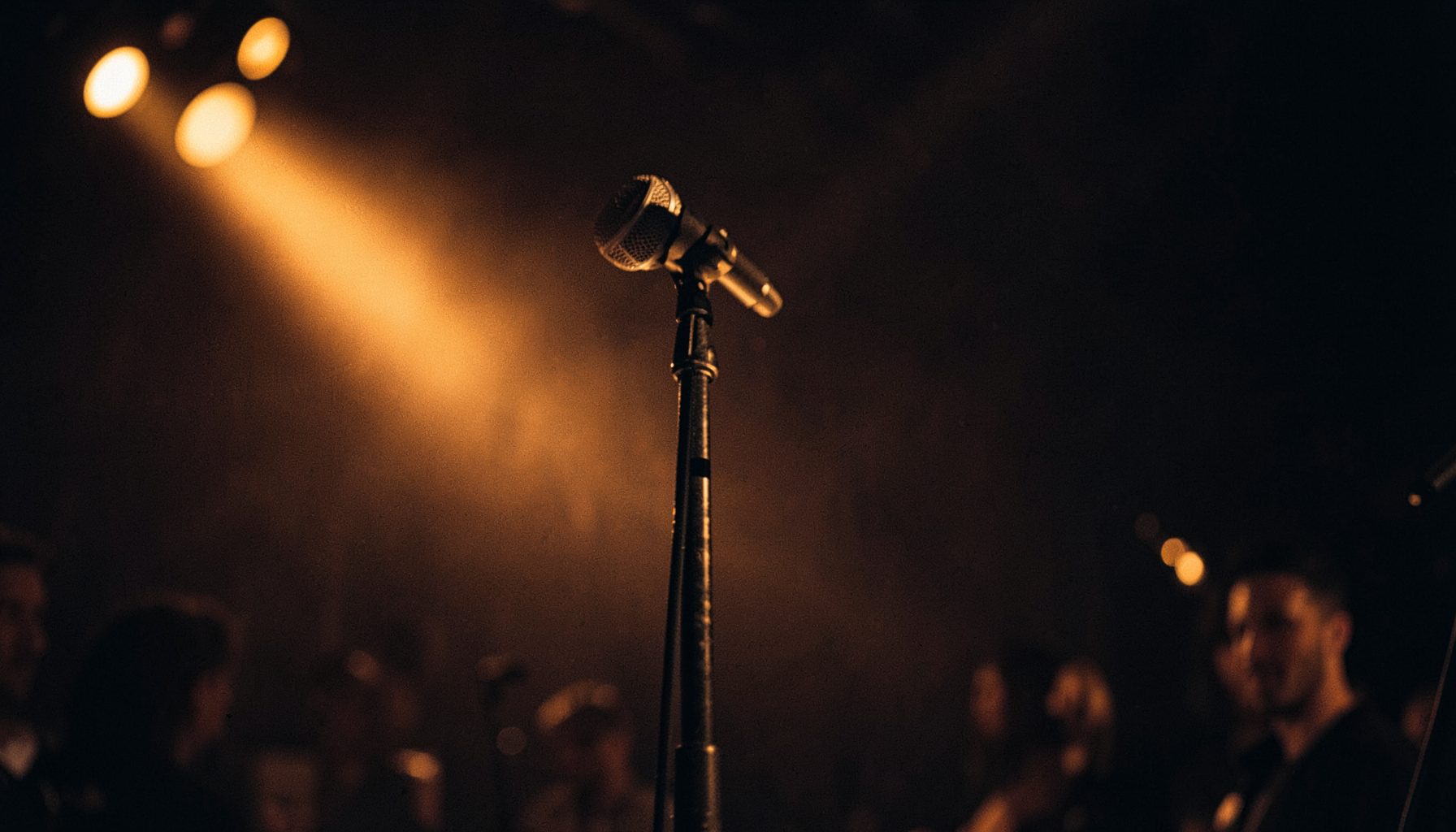 Warm, softly lit concert stage with a microphone stand and blurred amber stage lights against a dark background, evoking an intimate indie music venue atmosphere.